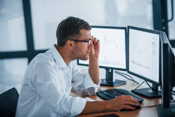 istock Young businessman in formal clothes is in office with multiple screens. Conception of exchange and money 1303446421