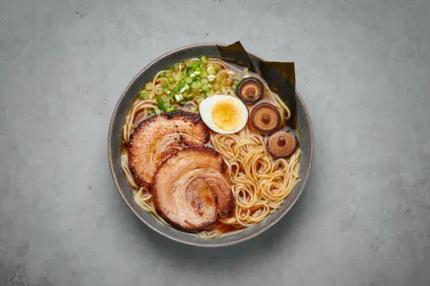 Photo of A Shoyu Ramen in gray bowl on concrete table top. Japanese cuisine meat noodle soup with chashu pork. Asian food. Top view