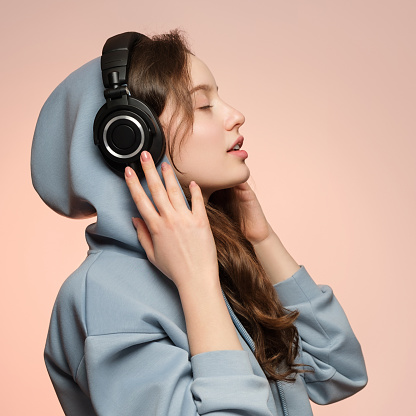 Girl in wireless headphones, face in profile on a soft pink background in a sizeless hoodie.
