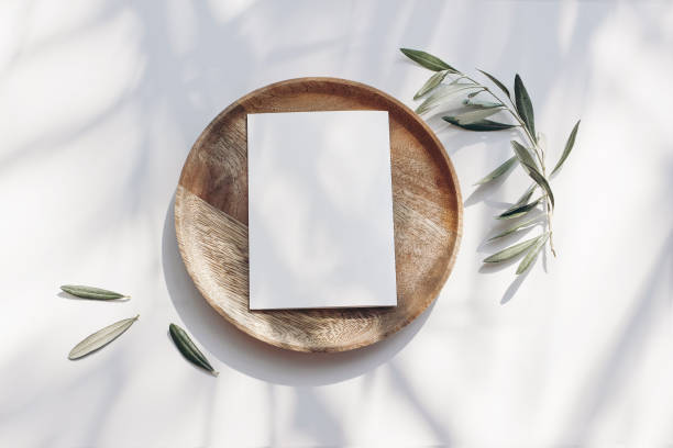 Summer wedding stationery mock-up scene. Blank greeting card, wooden plate, olive tree leaves and branches in sunlight. White table background with palm shadows. Feminine flat lay, top view. stock photo