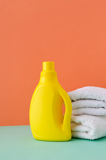 Yellow bottle of soft laundry detergent and white clean towels on the blue surface against orange background.Empty space
