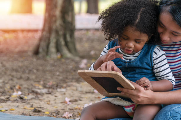 istock Close up young mother holding blackboards with her daughter while sitting on lawn learning something together in the park. Mixed race little curly hair girl sitting on knees of her mom. 1303460228