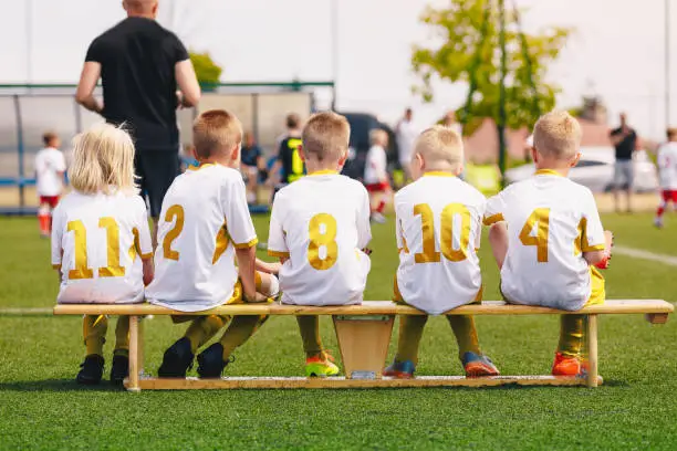 Photo of Group of Children in Soccer Jerseys with Golden Player Numbers. Football Team on Bench. Group of Young Boys with School Sports Coach Trainer. Youth Football Team Kicking Match in the Background