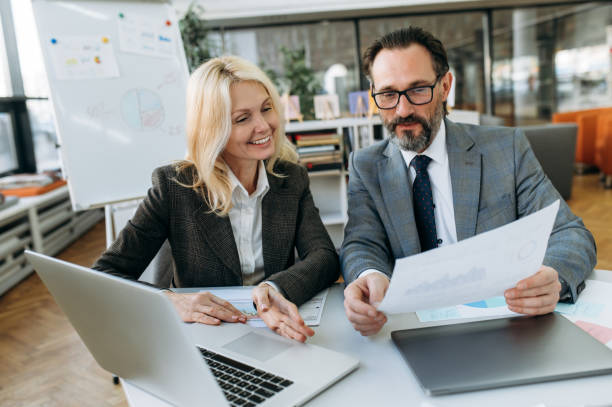 istock Middle aged friendly colleagues working together on project, sitting at the desk in office. Successful coworkers doing paperwork, discussing ideas for project, looking at financial graphs 1303467569