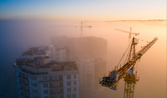 Construction site at dawn. Tower cranes over fog on the background of the morning sun. Drone view