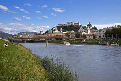 Salzburg, Land Salzburg, Austria, September 15, 2017: View from the embankment of the Salzach river to the Historic Centre of the City of Salzburg with the Hohensalzburg Fortress on the mountain in the background. Unidentified people rest on the bank of river and walk along the bridge and the river embankment.