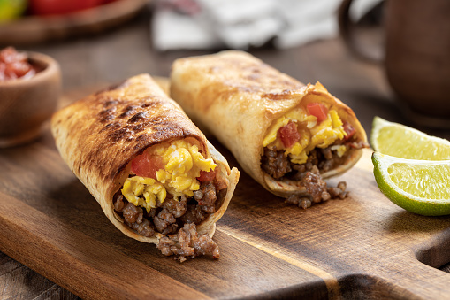 Two breakfast burritos with scrambled egg, sausage and tomato in a tortilla wrap