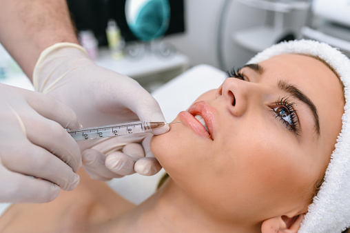 The doctor cosmetologist makes the Rejuvenating facial injections procedure for tightening and smoothing wrinkles on the face skin of a beautiful, young woman in a beauty salon. The hands of cosmetologist are close-ups that inject hyaluronic acid into the lips of the woman.