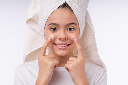 Cropped shot of a little teen girl in spa towel applying face cream on her cheeks isolated over white background