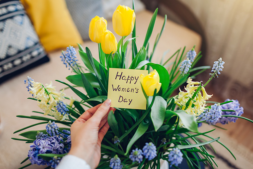Happy Womens Day, 8 March gift. Woman holds greeting card with blooming spring yellow blue flowers at home. Present for holiday with 2021 colors