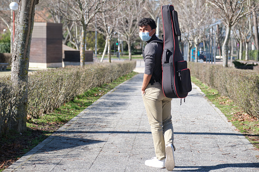 Latin young man wearing protective face mask carrying a guitar in a guitar case on a city street. University campus.