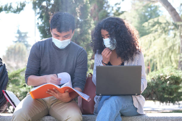 istock Two latin students wearing protective face mask studying together sitting on a bench outdoor. New normal at university campus. 1303494317