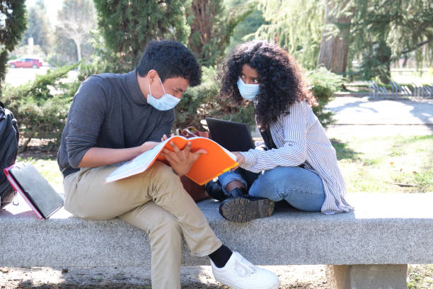 istock Two latin students wearing protective face mask studying together sitting on a bench outdoor. New normal at university campus. 1303494366