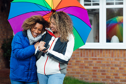 Sisters standing outdoors in the rain laughing with each other holding a multicoloured umbrella above their heads.