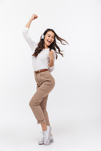 Full length portrait of a happy asian businesswoman listening to music with headphones while dancing isolated over white background