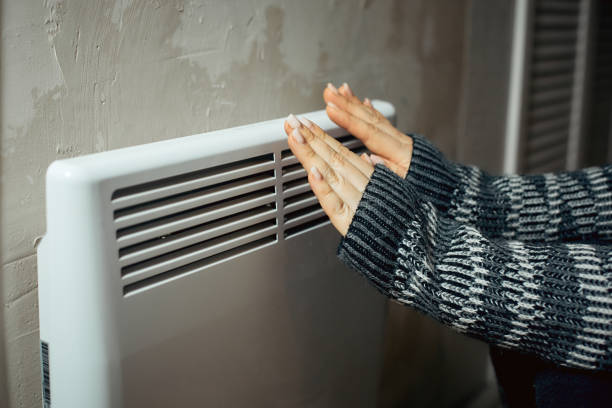 a woman warms her hands at the radiator in a cold house, problems with heating, heating the room with an electric convector stock photo