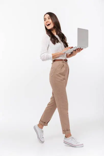 Full length portrait of a happy asian businesswoman holding laptop computer and looking away isolated over white background