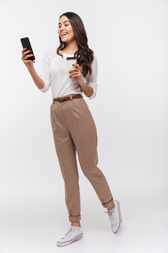 Full length portrait of a smiling asian businesswoman using mobile phone while holding cup of coffee to go isolated over white background