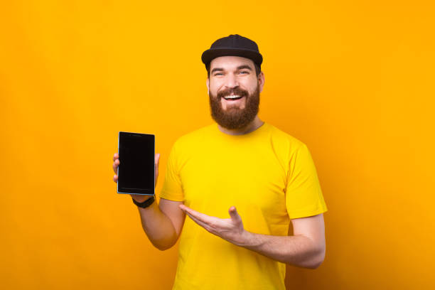istock Cheerful bearded young man showing tablet over yellow background 1303502669
