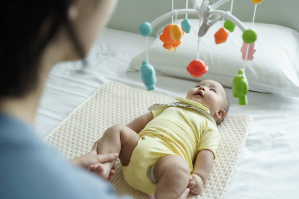 Asian Mother holding her son legs while baby boy lying on bed playing with mobile.