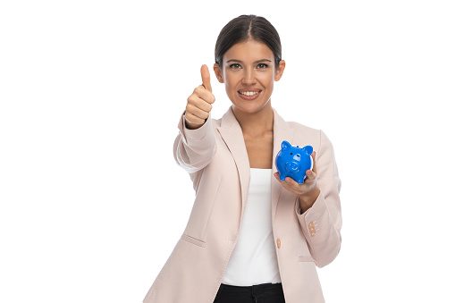 attractive brunette woman in pink jacket making thumbs up gesture, smiling and showing piggy bank on white background in studio