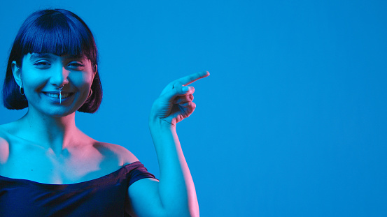 Young mixed race brunette cheerful woman point with her finger isolated on blue neon copyspace