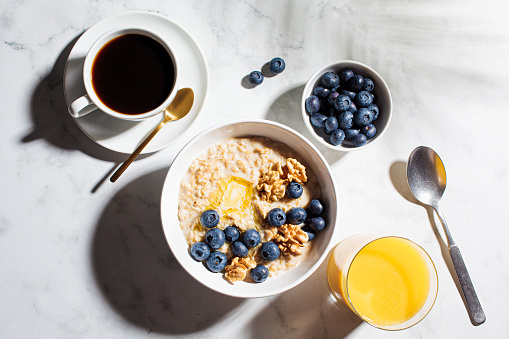 Breakfast concept. Oatmeal with berries and nuts, a cup of coffee and a glass of orange juice on a white marble background.