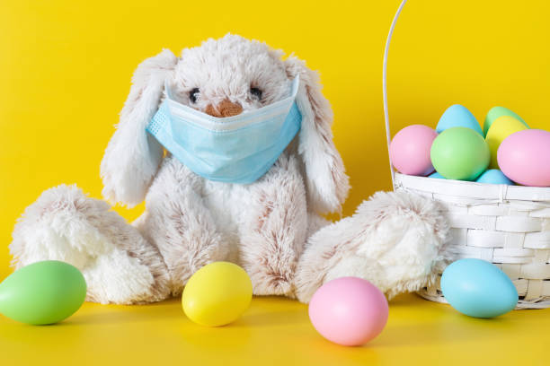 istock Easter bunny in mask and colored eggs in basket on yellow background. Ester holiday during corona virus concept. 1303529744