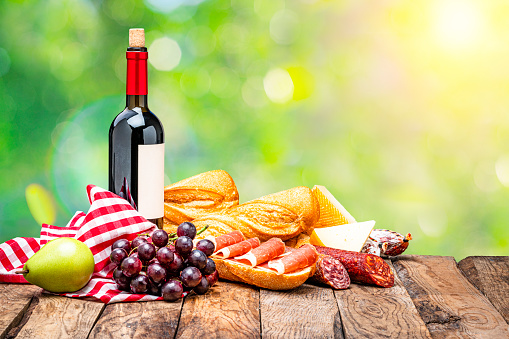 Picnic food: wooden table with delicious food shot outdoors against defocused lush foliage. The composition includes bread, grapes, various cheeses, a Iberico ham sandwich, chorizo and a red wine bottle. Copy space available at the right. High resolution 42Mp studio digital capture taken with Sony A7rII and Sony FE 90mm f2.8 macro G OSS lens