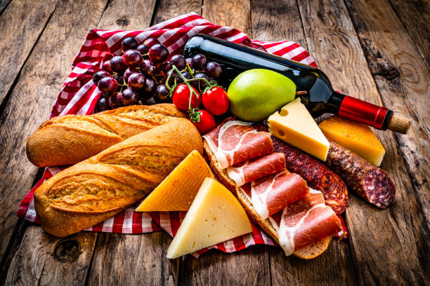 Picnic food: high angle view wooden table with delicious food grouped at the center of the frame. The composition includes bread, grapes, various cheeses, a Iberico ham sandwich, chorizo and a red wine bottle. High resolution 42Mp studio digital capture taken with Sony A7rII and Sony FE 90mm f2.8 macro G OSS lens