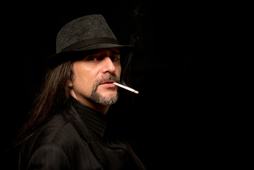 Portrait of a man smoking cigarette, wearing black hat and black suit.