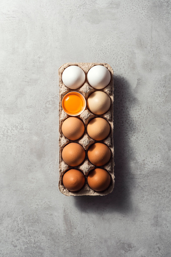 Box of color gradient brown eggs with one open egg with yolk, rectangular egg carton on grey background. Easter eggs. Shot from directly above.