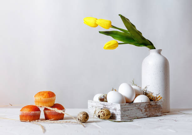 istock Three cupcakes on a light table. White chicken eggs and quail eggs in a wooden box with straw on a white light background. Tulip flower in a vase in the background. 1303546777
