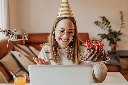A young woman is in the living room, she is celebrating birthday via video call on a laptop