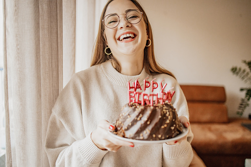 A young woman is at home, she is smiling and holding a birthday cake