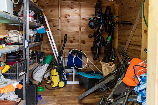 Suburban home wooden storage utility unit shed with miscellaneous stuff on shelves, bikes, exercise machine, ladder, garden tools and equipment. Messy and chaos at house yard barn. Organization order.