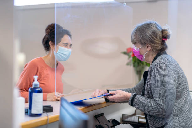Receptionist welcoming young mixed race female patient A young mixed race Pacific Islander woman wearing a protective face mask checks in at a reception desk outfitted with an acrylic glass sneeze guard before a medical or dental appointment during the COVID-19 pandemic. The receptionist is helping the patient with a registration form and is also wearing a protective face mask. registration form photos stock pictures, royalty-free photos & images