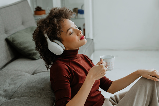 A pretty Afro woman sitting on the living room floor listening to music, while enjoying a cup of coffee.