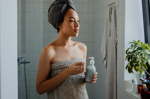 A gorgeous young woman standing in the bathroom after a shower, wrapped in a towel and holding a bottle of liquid soap.