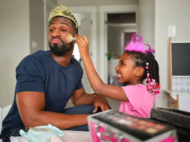 An African American father and daughter playing dress up and putting on makeup n their home.