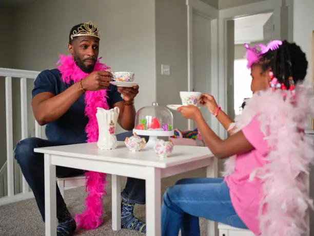An African American father and daughter playing and pretending to have a tea party in their home.