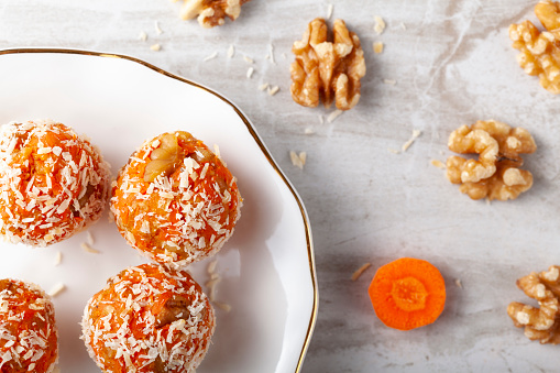 A flat lay of kitchen countertop with a flat porcelain plate. Homemade fresh carrot bliss balls (mixture of cookie crumbles, walnut and carrot puree) coated with coconut shred are served on the plate.