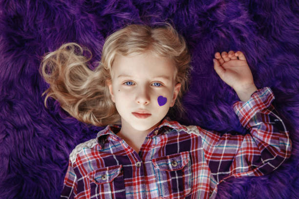 dia internacional mundial de conscientização da doença da epilepsia. linda garota branca loira com pequeno coração de papel violeta roxo na bochecha deitado no tapete roxo fofo em casa. veja de cima. - epilepsy - fotografias e filmes do acervo
