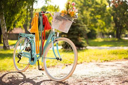 Beautiful turquoise bicycle parked in a park. Different color fashion bags are hanging on a governor. Summer flowers in bicycle white basket.