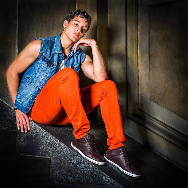 A young handsome American college student is wearing Denim sleeveless jacket, red pants, brown leather shoes, sitting by stairs inside an office building, a hand touching face. A fine art fashion color photography\