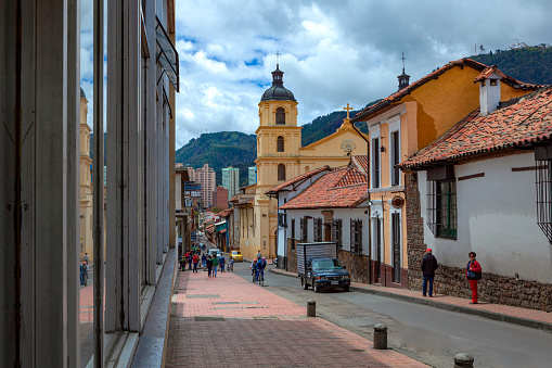 Bogota, Colombia - July 20, 2016: Looking downhill at one of the Baroque Towers of the Church of Our Lady of Candelaria, in the South American capital city of Bogota in Colombia. The Church was built between 1686 and 1703 by the Catholic Augustinian Order.  Local Colombian people walk through the narrow streets of the historical La Candelaria district of Bogotá, the Andean capital city of the South American country of Colombia. It is a holiday in the Country and there is hardly any traffic around. It is in this area that the Spanish Conquistador, Gonzalo Jiménez de Quesada founded the city in 1538. Many of the walls in this area are painted with street art, legends of the pre Colombian era, or just in the vibrant colours of Colombia. The sky is overcast: it will probably rain shortly. Photo shot on a cloudy morning; horizontal format. Copy space.