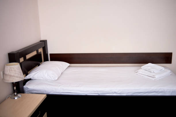 istock Hotel bedroom interior with empty single bed with wooden headboard and bedside table near beige wall, copy space. White sheet, soft pillow and towels on bed 1303589680