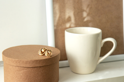 Still life. Cup of coffee, blank beige poster frame, craft round box and beautiful gold earrings on the white wall. Side view