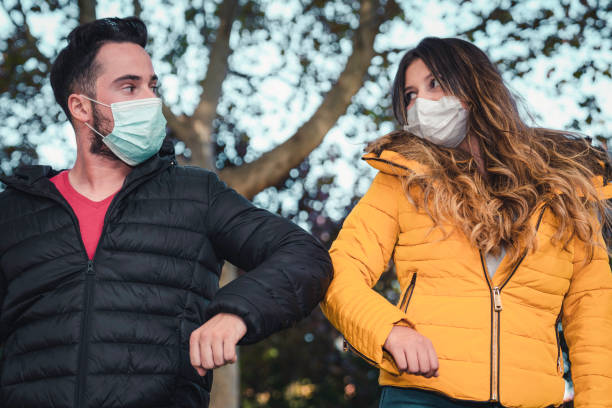 istock Young beautiful couple in protective masks greeting each other with elbow touch gesture in park 1303595825