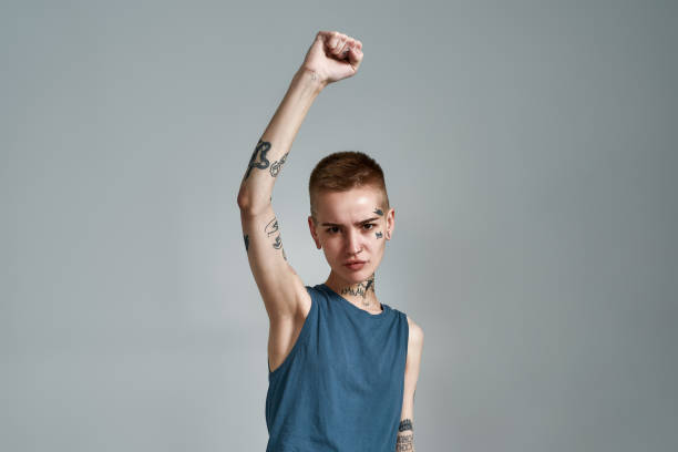 Disgruntled tattooed young caucasian woman protester with piercing looking at camera while posing with raised arm isolated over gray background